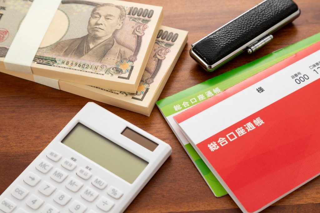 Stacks of Japanese yen bundles, a calculator, a pen, and red/green accounting ledgers on a wooden desk.