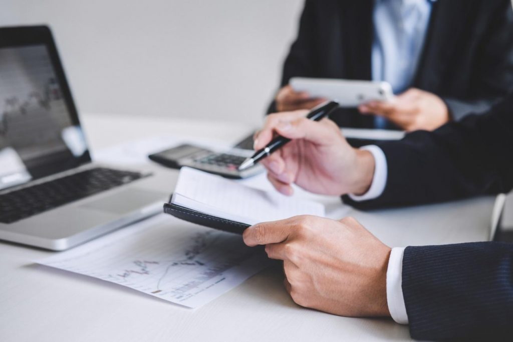 Two professionals in suits review documents on a desk with a notebook, calculator, tablet, and laptop nearby.