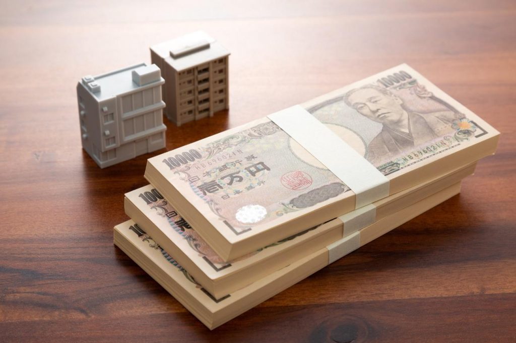 Stack of Japanese 10,000 yen notes bound with white bands beside miniature model buildings on a wooden table.