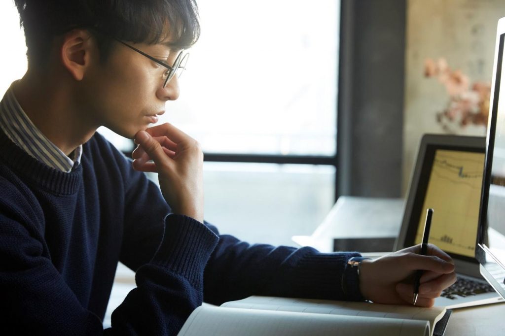 Student in glasses writes in a notebook, with a laptop nearby in a bright study space, appears to be thinking.
