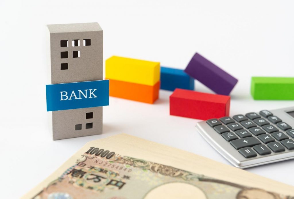 Toy bank building with a blue 'BANK' banner, colorful blocks, a calculator, and a 10,000 yen note on a white background.