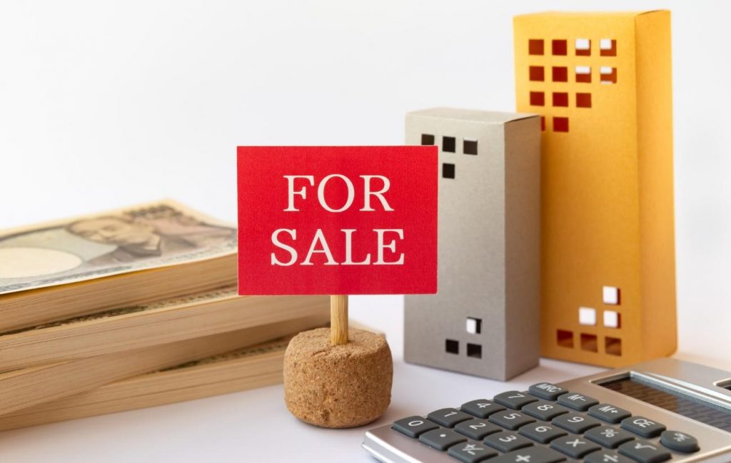 For sale sign with miniature buildings, cash, and a calculator illustrating real estate pricing/availability.