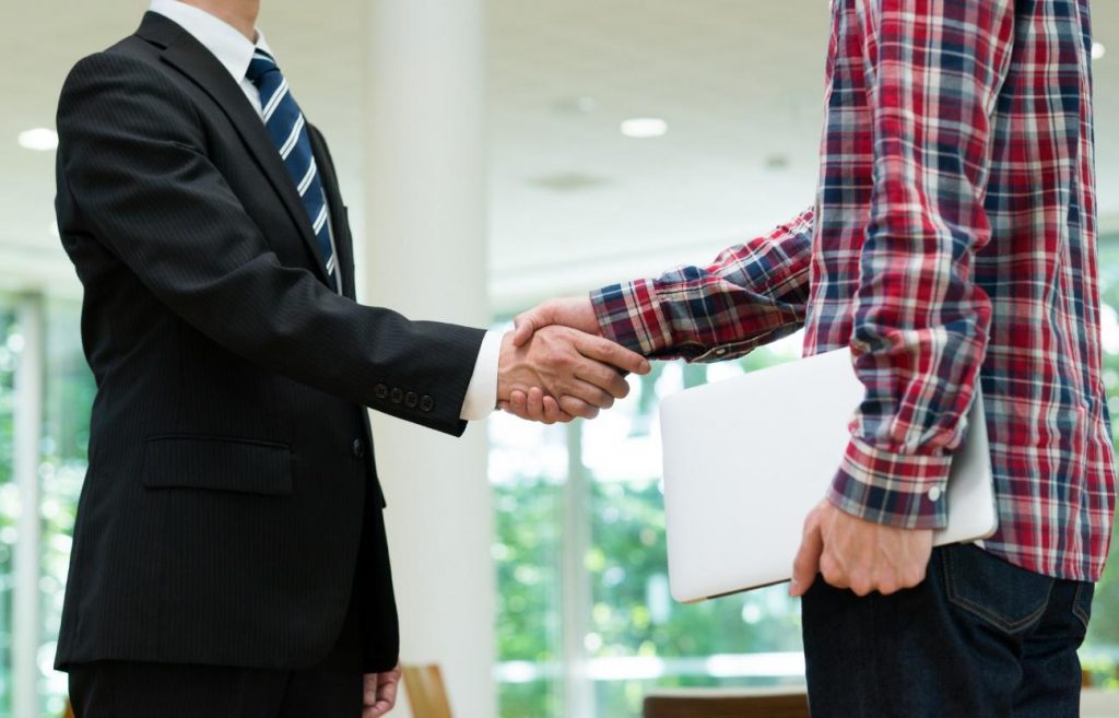 Two people shake hands in a bright office; one in a suit, the other in a plaid shirt, with a laptop nearby.