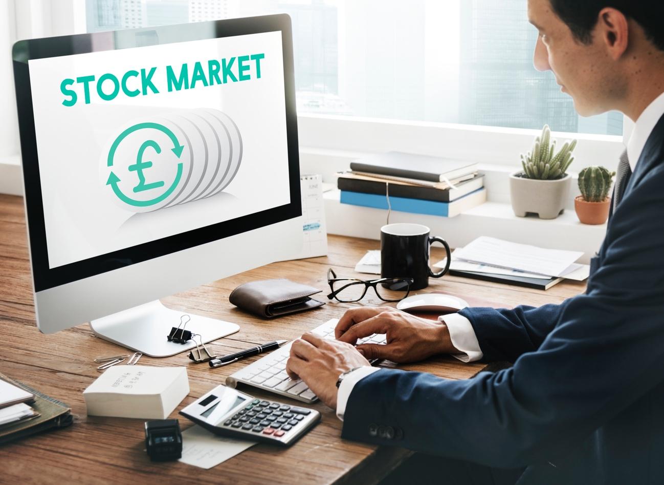 Businessperson typing at a desk with a computer monitor displaying a stock market graphic and a pound symbol in teal.