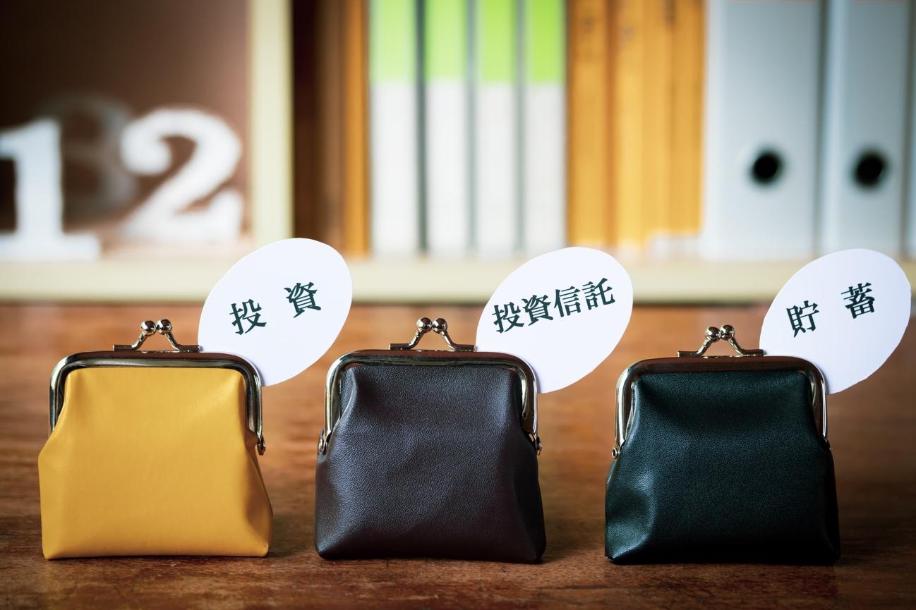 Three small coin purses in yellow, black, and dark blue sit on a wooden surface, each with a white circle label displaying Japanese terms for investment, mutual funds, and savings (投資, 投資信託, 貯蓄).