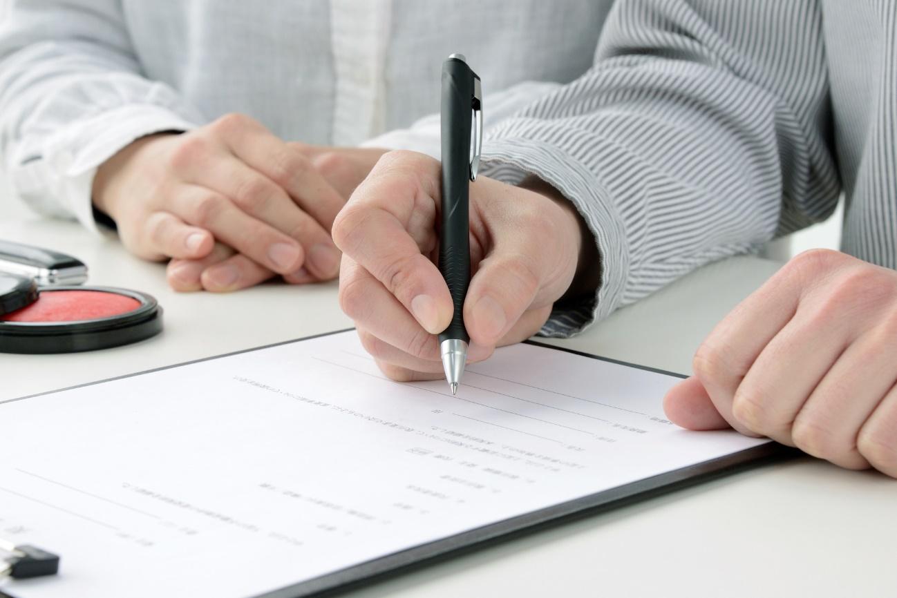 Person signs a form with a black pen at a white desk; a makeup compact is nearby in the background for context.