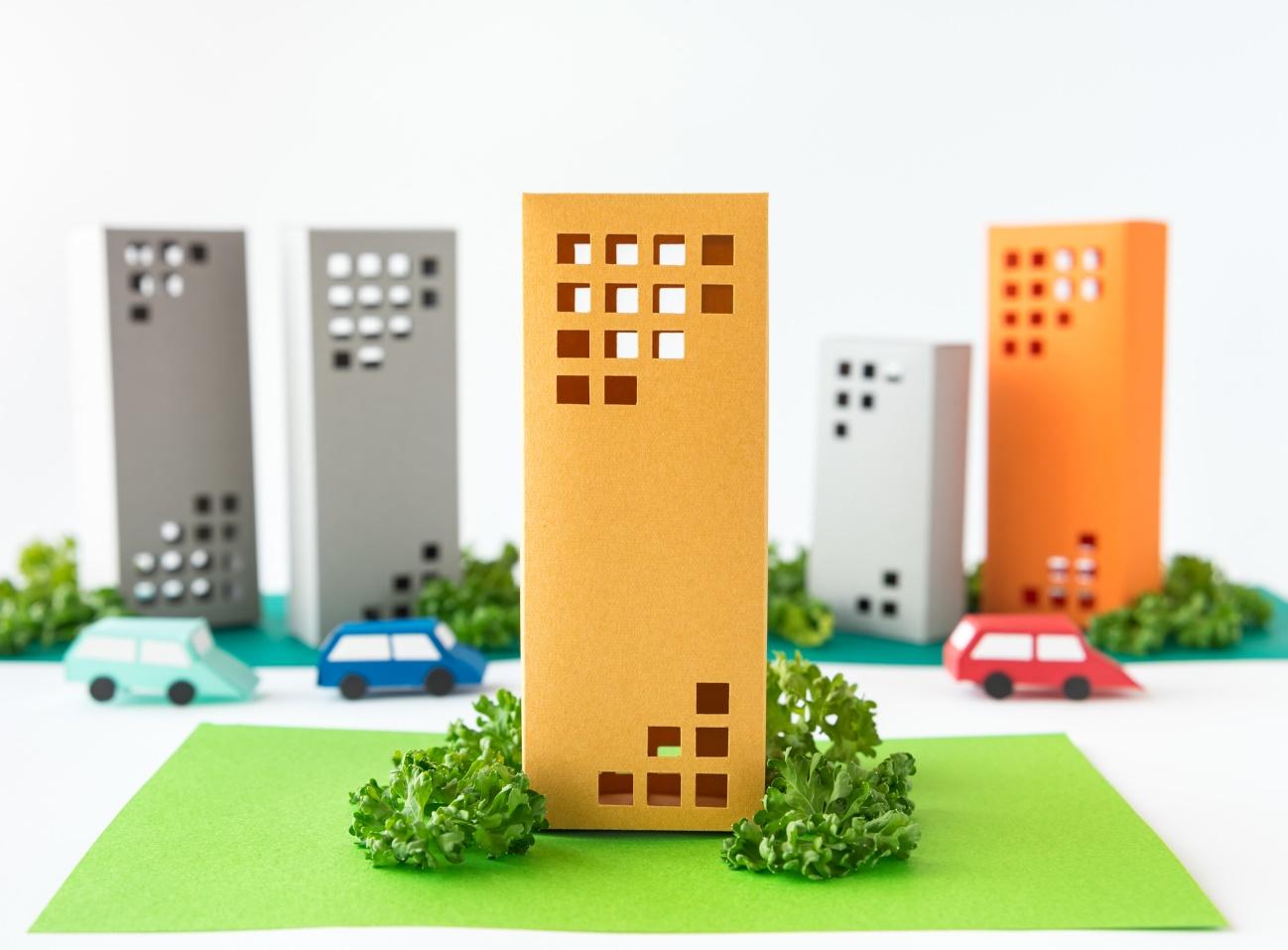 Paper city model with a central brown-orange building in the foreground, surrounded by other colorful buildings and toy cars on green ground.