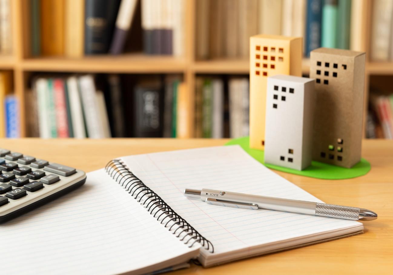 Notebook and calculator on a wooden desk with paper-model buildings beside a bright green base, bookshelves blurred in the background.