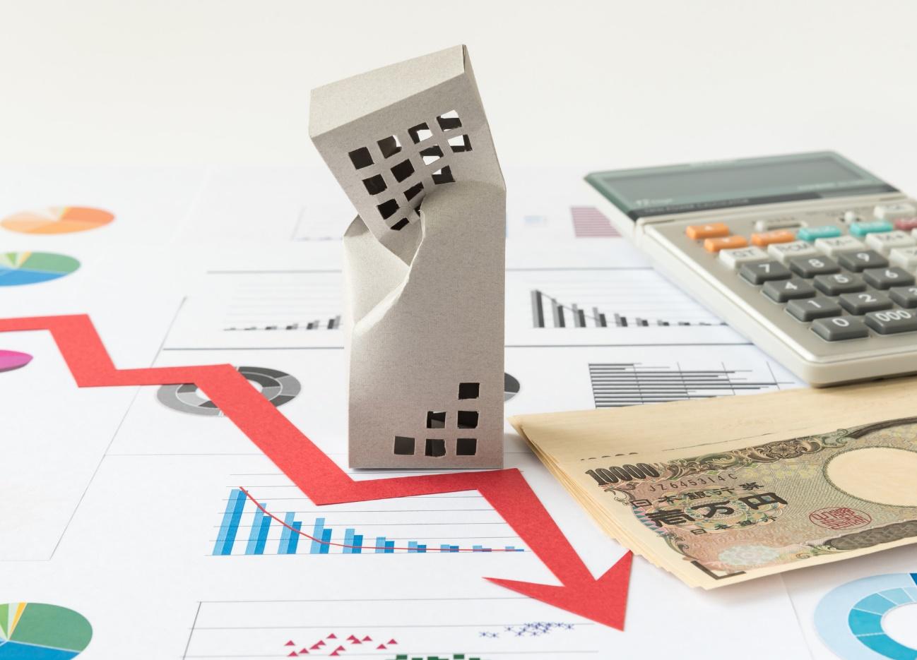 Cardboard house model placed on financial charts with a red downward arrow, calculator, and yen banknotes, signaling a housing market decline,