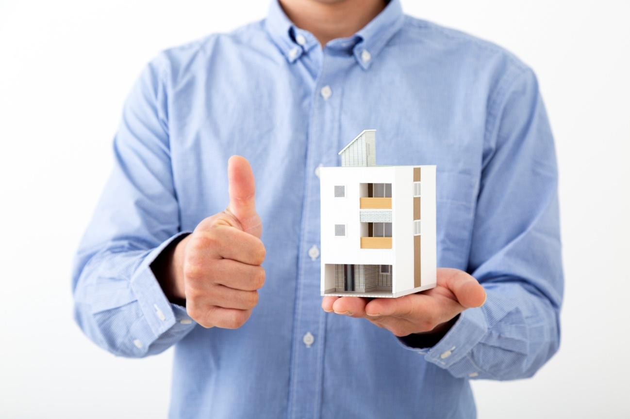Person in a blue shirt holding a small modern building model and giving a thumbs-up gesture.