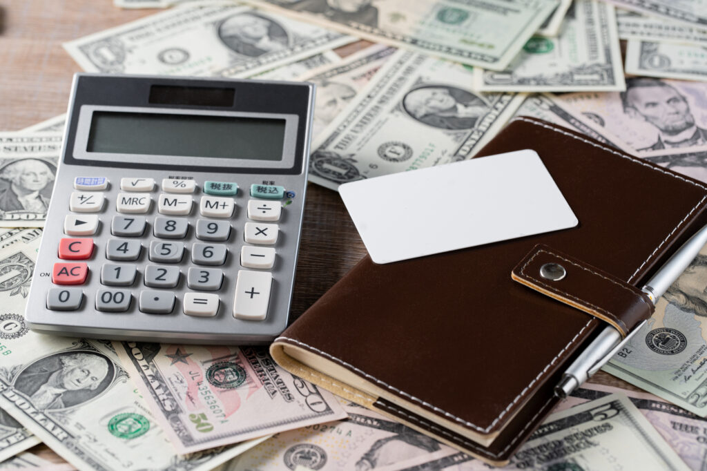 Calculator atop scattered dollar bills with a brown leather wallet and blank card nearby, suggesting finances or budgeting.