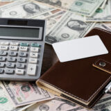 Calculator atop scattered dollar bills with a brown leather wallet and blank card nearby, suggesting finances or budgeting.
