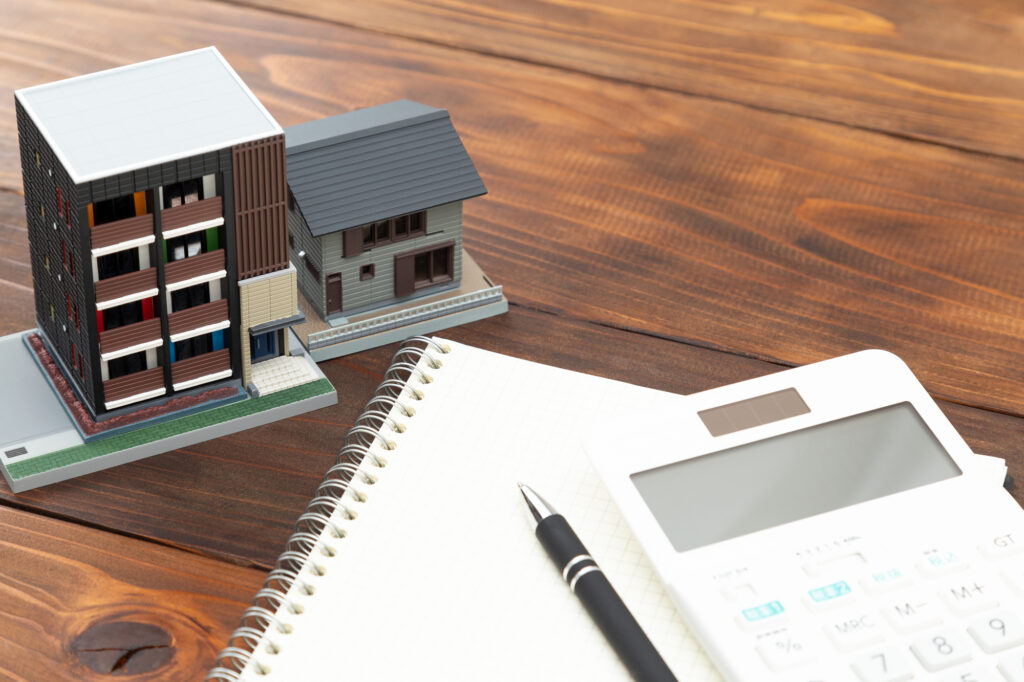 Miniature building models beside a spiral notebook, pen, and calculator on a wooden desk, suggesting architecture planning.