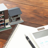 Miniature building models beside a spiral notebook, pen, and calculator on a wooden desk, suggesting architecture planning.