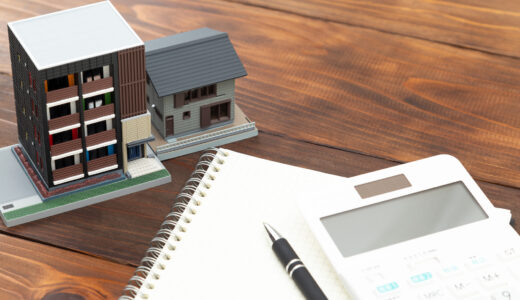 Miniature building models beside a spiral notebook, pen, and calculator on a wooden desk, suggesting architecture planning.