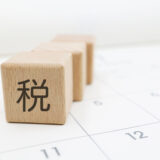 Wooden blocks arranged on a calendar grid, front block showing the Kanji '税' (tax); a gray hash symbol floats to the left.