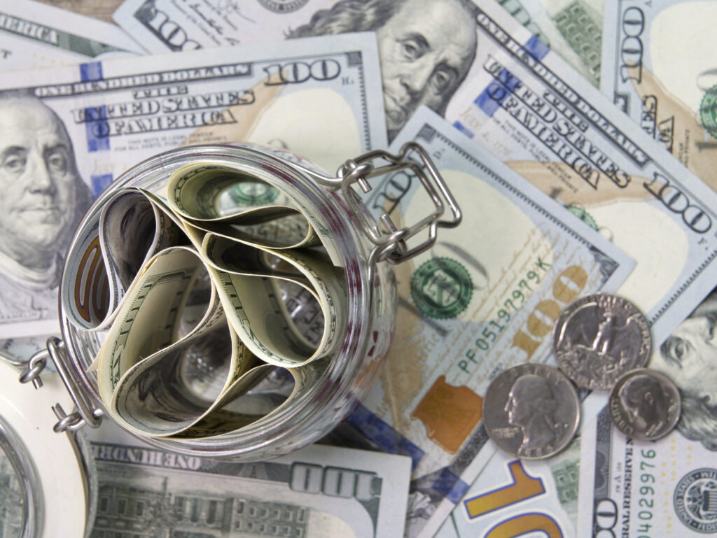 Close-up of a glass jar filled with rolled dollar bills and coins on a spread of cash behind it, conveying money storage/ savings.