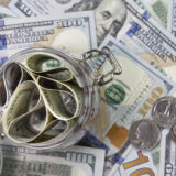Close-up of a glass jar filled with rolled dollar bills and coins on a spread of cash behind it, conveying money storage/ savings.