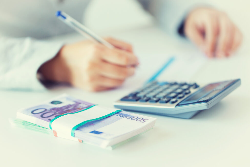 Person writing with a pen beside a calculator and a stack of euro banknotes, suggesting budgeting or accounting (blurry).