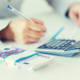 Person writing with a pen beside a calculator and a stack of euro banknotes, suggesting budgeting or accounting (blurry).
