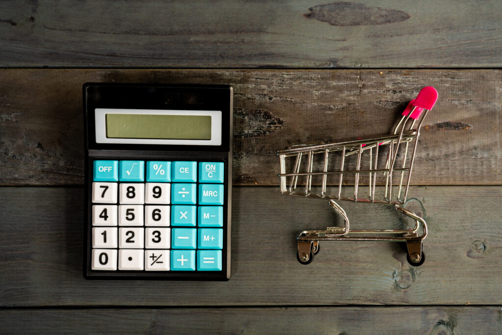Black calculator with blue and white numeric keys sits on a dark wooden surface beside a small metal shopping cart on a wooden tabletop, display blank.