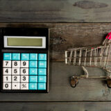 Black calculator with blue and white numeric keys sits on a dark wooden surface beside a small metal shopping cart on a wooden tabletop, display blank.