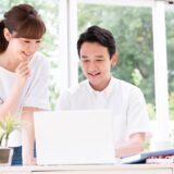 Two colleagues, a woman and a man, smile while looking at a laptop on a bright desk near a window with greenery outside.