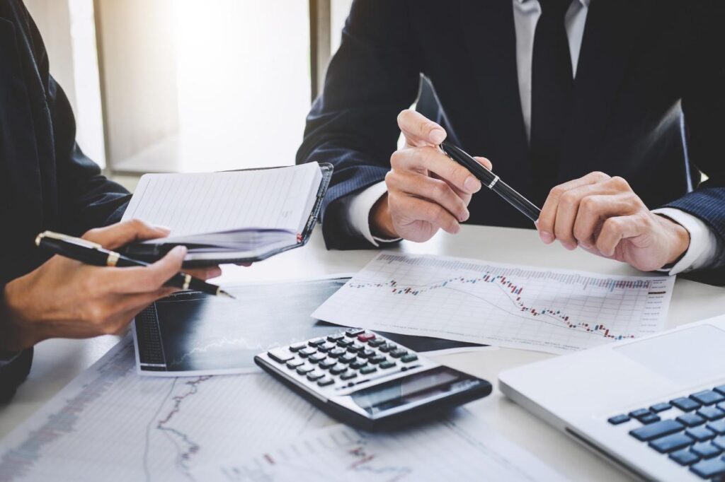Two professionals in business attire review financial reports and charts on a white desk, with a calculator, notebook, and laptop nearby.
