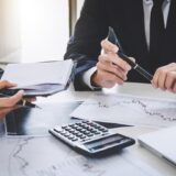 Two professionals in business attire review financial reports and charts on a white desk, with a calculator, notebook, and laptop nearby.