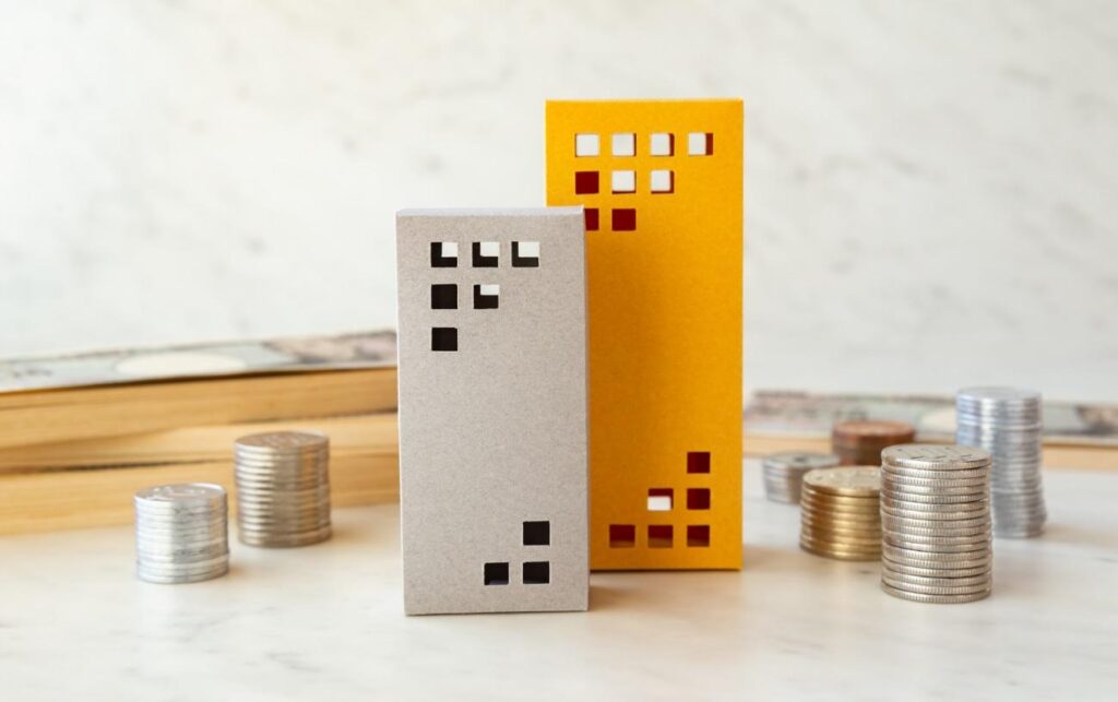 Two decorative cardboard houses with square window cutouts, placed on a light surface near stacks of coins—real estate themed.