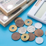 Stacks of Japanese yen banknotes with coins and a white calculator on a blue surface.