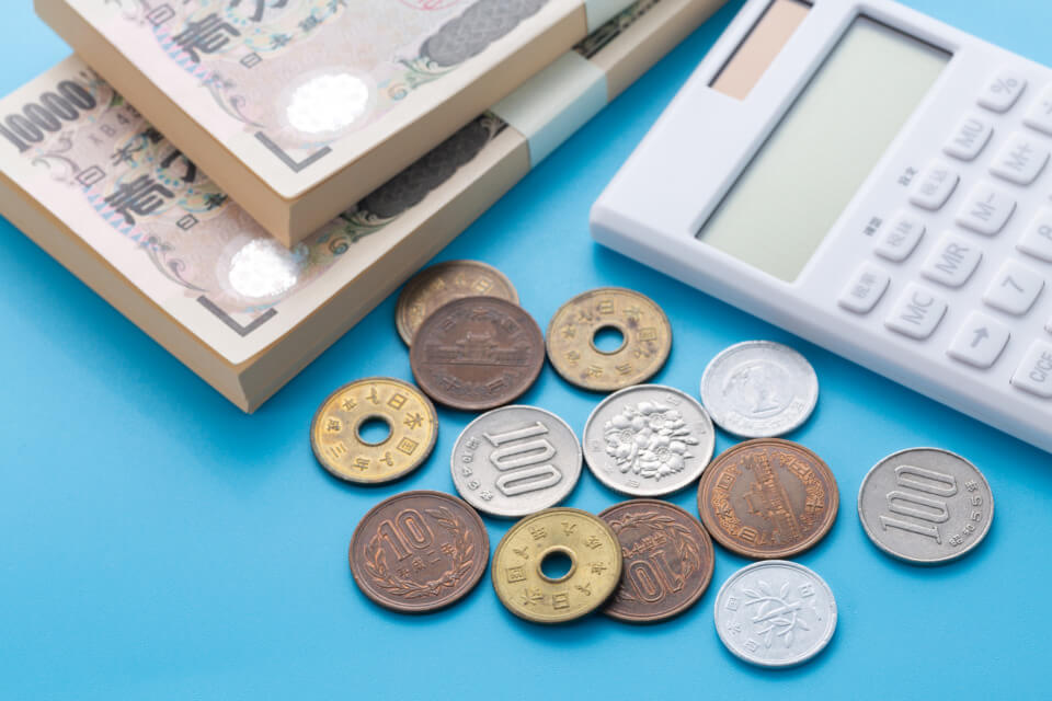 Stacks of Japanese yen banknotes with coins and a white calculator on a blue surface.