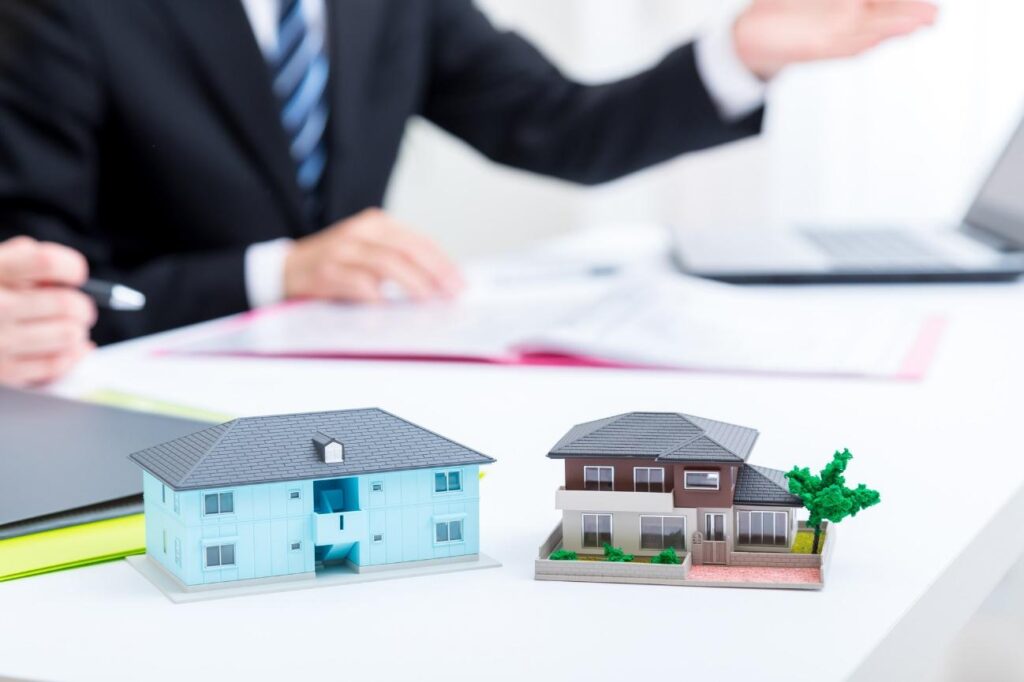 Two miniature house models on a desk with a blurred businessman in a suit in the background, suggesting real estate planning.