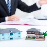 Two miniature house models on a desk with a blurred businessman in a suit in the background, suggesting real estate planning.