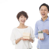 Two smiling adults, a woman holding a small wooden house and a man in a blue shirt holding a calculator, posing happily.