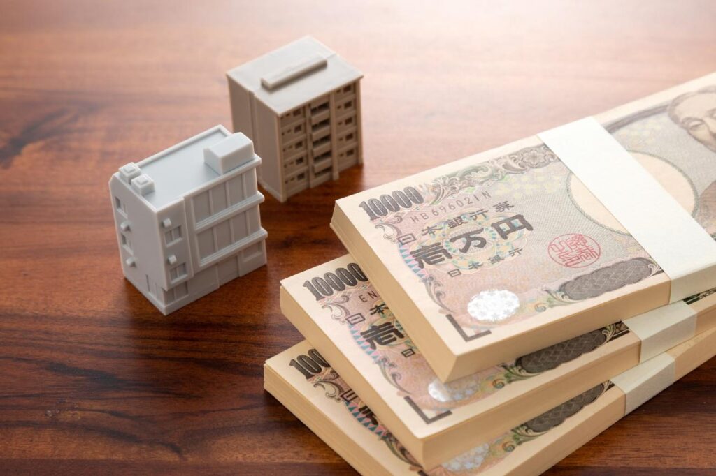 Stacks of 10,000 yen banknotes on a wooden table with white toy buildings, suggesting real estate or investment theme.