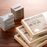 Stacks of 10,000 yen banknotes on a wooden table with white toy buildings, suggesting real estate or investment theme.