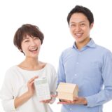 Two smiling adults hold a calculator and a small wooden house model, suggesting home budgeting or mortgage planning.