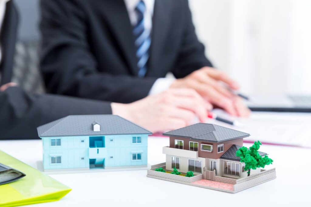 Scale models of a blue and a brown house on a white desk, with blurred hands of two businesspeople working in the background