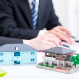 Scale models of a blue and a brown house on a white desk, with blurred hands of two businesspeople working in the background