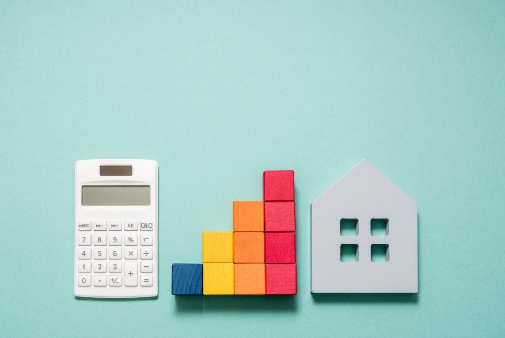White calculator beside a multicolored wooden block bar chart and a gray house silhouette on a teal background, indicating budgeting for housing.