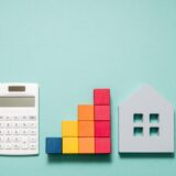 White calculator beside a multicolored wooden block bar chart and a gray house silhouette on a teal background, indicating budgeting for housing.