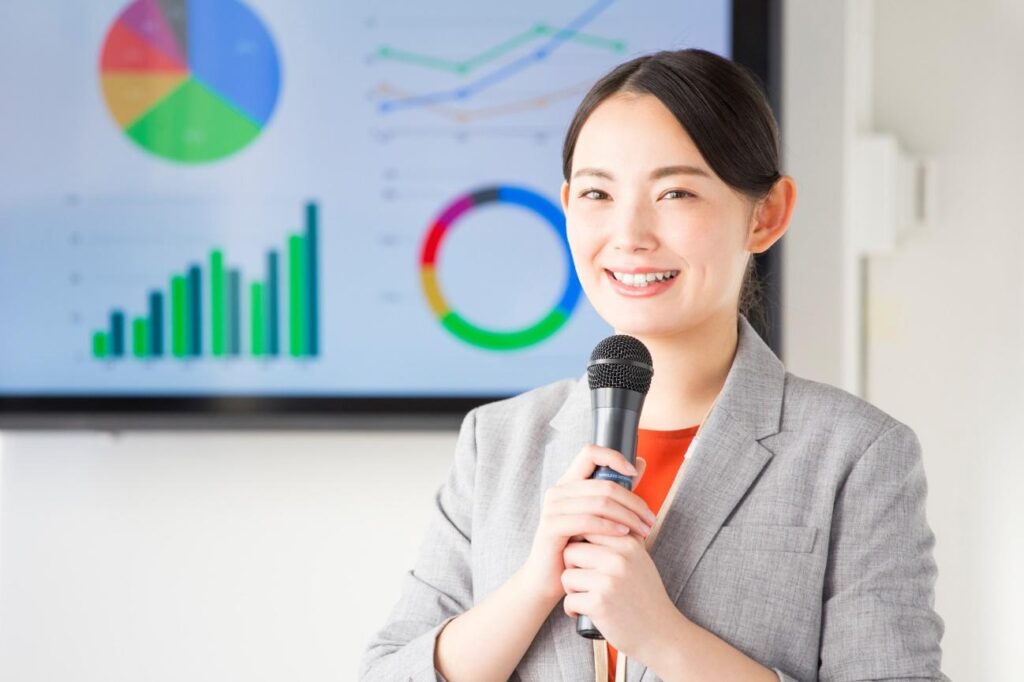 Smiling Asian woman in a gray blazer holding a microphone during a presentation, with colorful charts on a screen behind her.