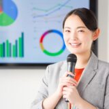 Smiling Asian woman in a gray blazer holding a microphone during a presentation, with colorful charts on a screen behind her.