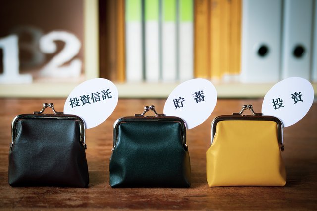 Three small coin purses on a wooden surface, each with a speech bubble label reading 投資信託, 貯蓄, 投資, against a blurred bookshelf background.