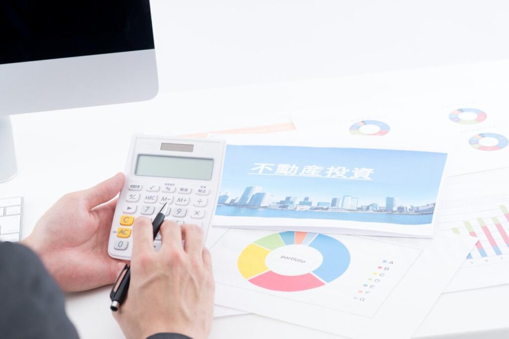 Person uses a calculator at a desk with colorful financial charts and a card that reads '不動産投資' (real estate investment).