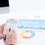 Person uses a calculator at a desk with colorful financial charts and a card that reads '不動産投資' (real estate investment).