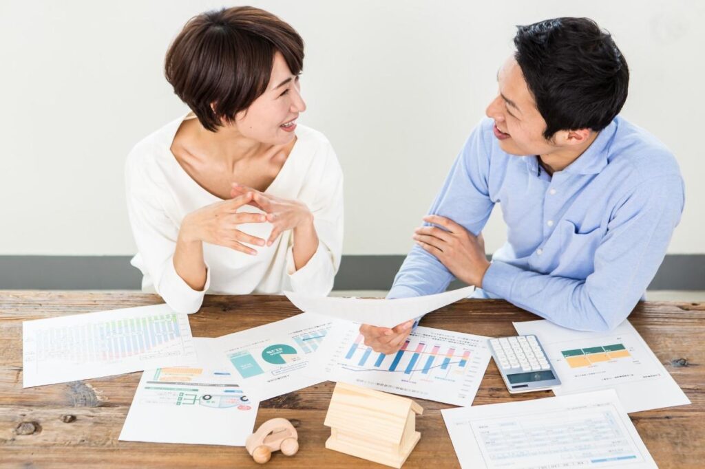 Two coworkers smiling while discussing charts and reports spread on a wooden table.