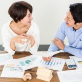 Two coworkers smiling while discussing charts and reports spread on a wooden table.