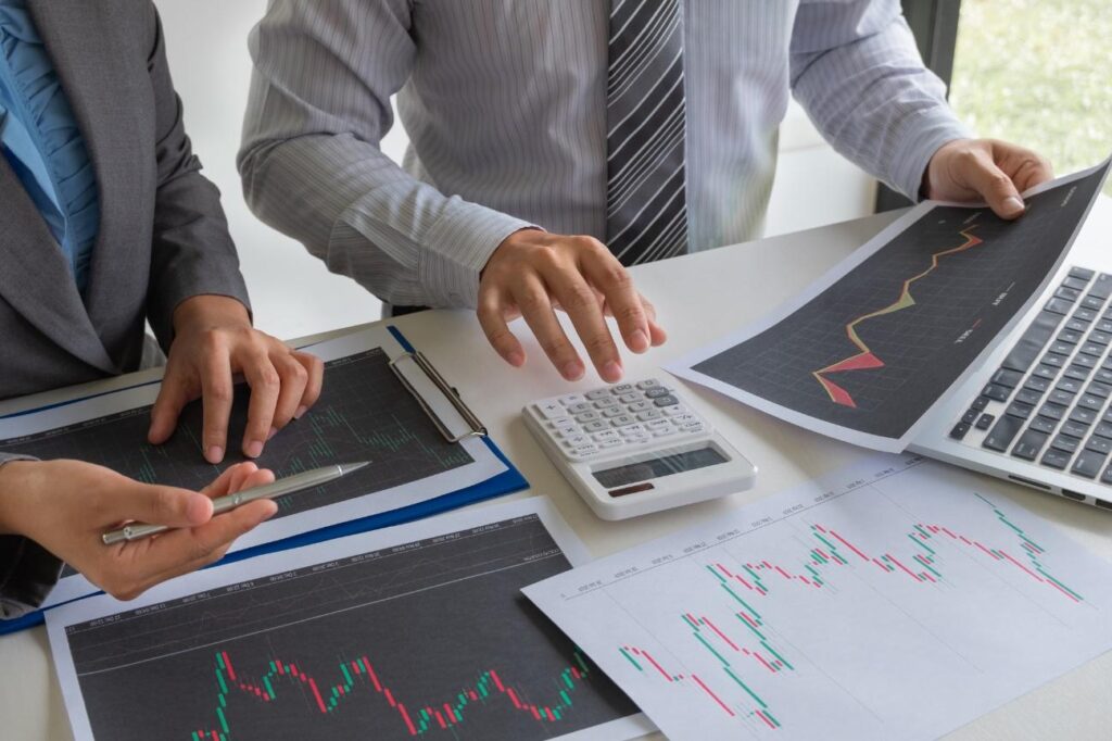 Two business professionals review financial charts: printed line charts, a calculator, and a laptop on a desk |in a meeting setting|, concise and informative for finance context.